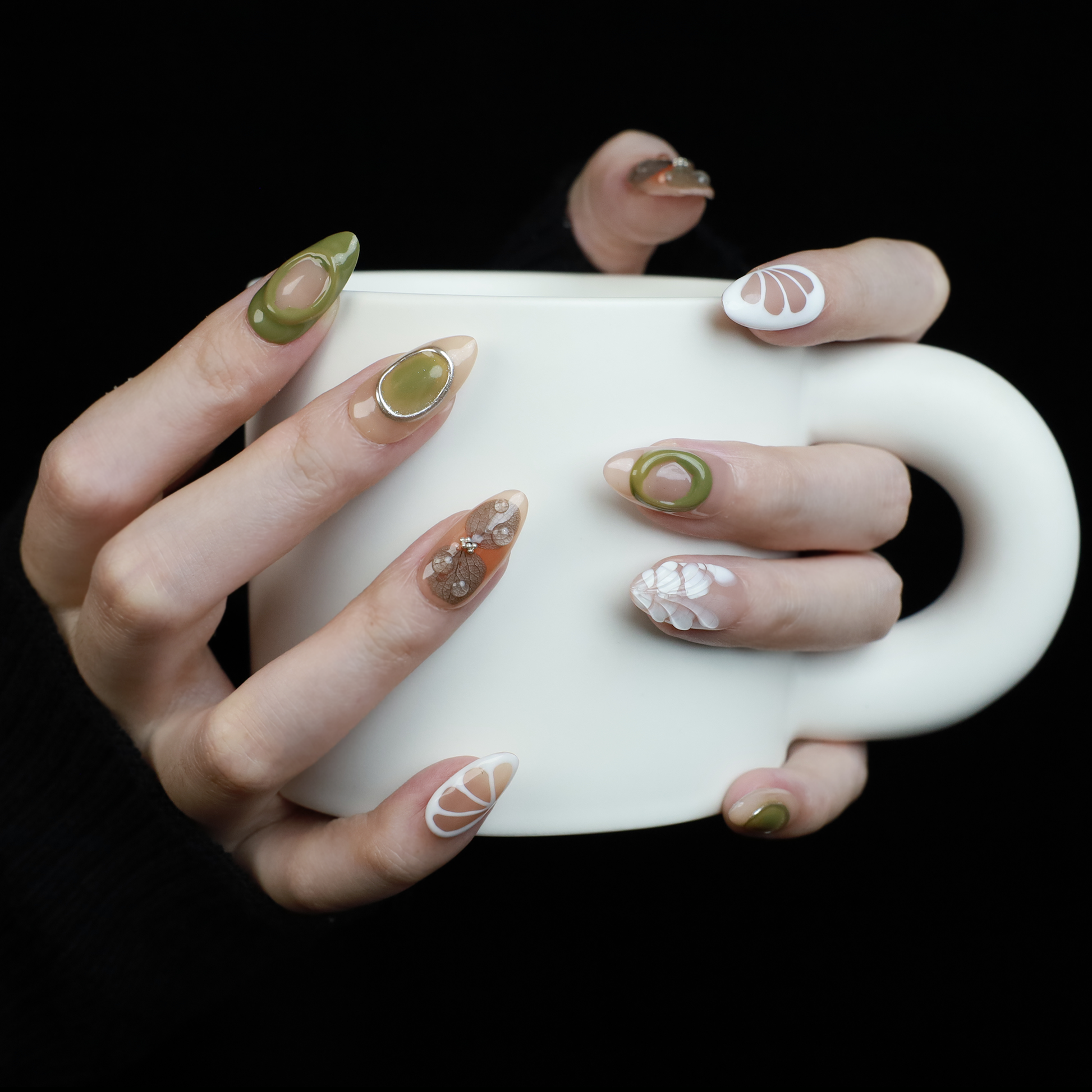 A hand in a black sleeve displays the Leaf and Organic design Press-On Nails by We Do Nails, featuring long acrylic nails with white abstract patterns, green accents, and clear 3D embellishments that create a nature-inspired look against the dark background.