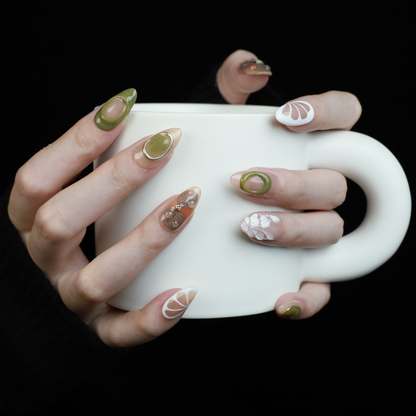 A hand in a black sleeve displays the Leaf and Organic design Press-On Nails by We Do Nails, featuring long acrylic nails with white abstract patterns, green accents, and clear 3D embellishments that create a nature-inspired look against the dark background.