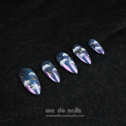 A hand showcases Starry Night Press-On Nails with long, glittery tips featuring a cloud and starry night design adorned with small gems. The person wears a black sleeve. Text below reads: We Do Nails with a website link.