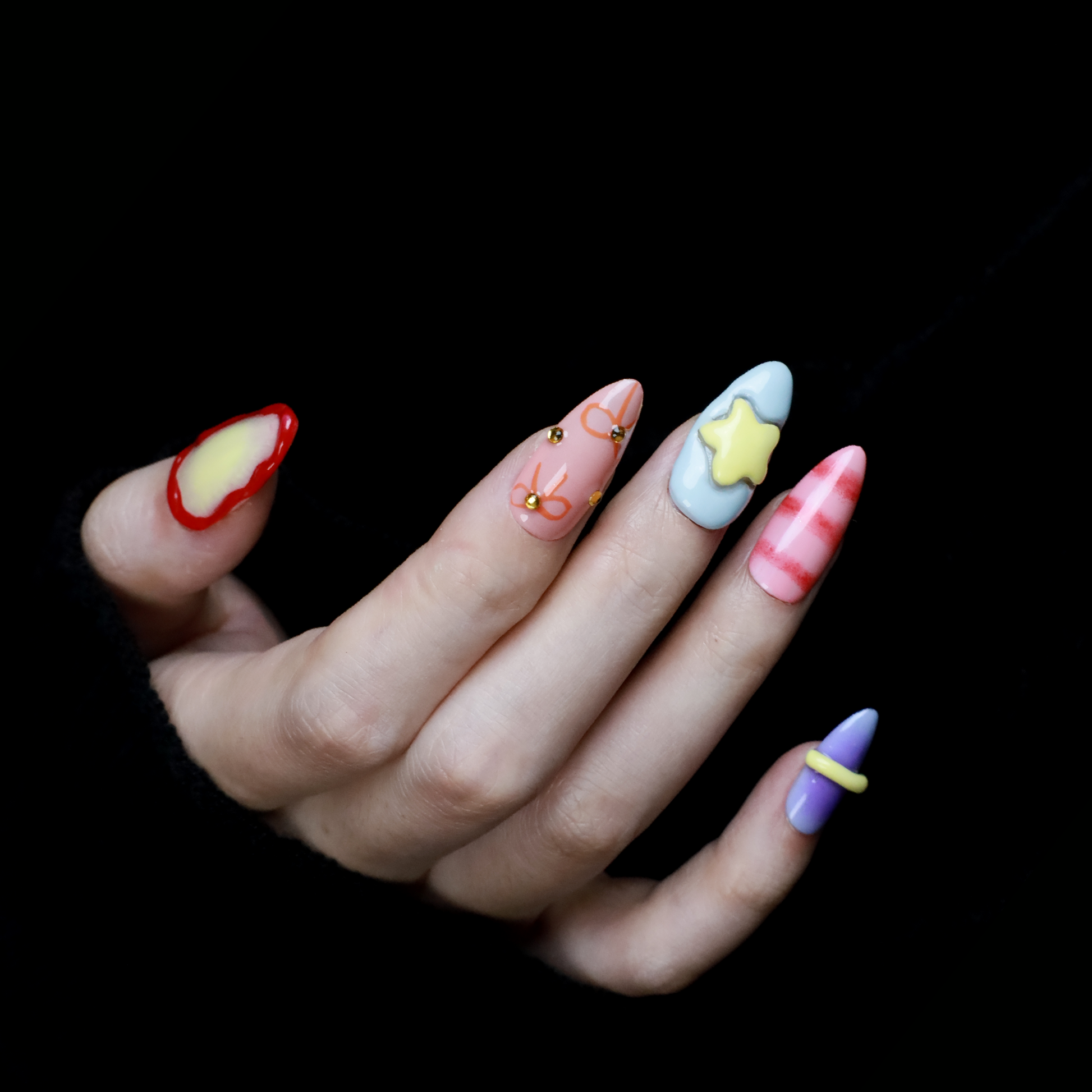A hand with We Do Nails Candyland Dream Press-On Nails displays playful designs. Features include pastel yellow plaid, clear nails with bows and rhinestones, pink nails with a bow, shiny red with rhinestones, and glossy rust-orange—all set against a dark backdrop.
