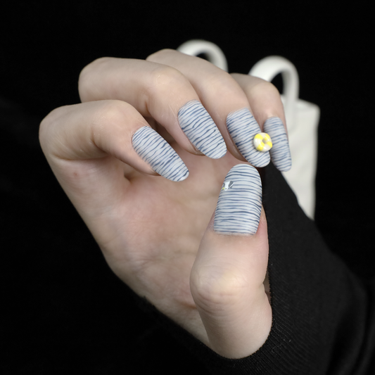 A hand with long nails showcases We Do Nails Nautical Navy Blue Stripes press-on nails, featuring a nautical blue and black wavy design. One nail is adorned with a 3D yellow flower, another with a small gem. A black sleeve adds elegance to the hand-painted look.