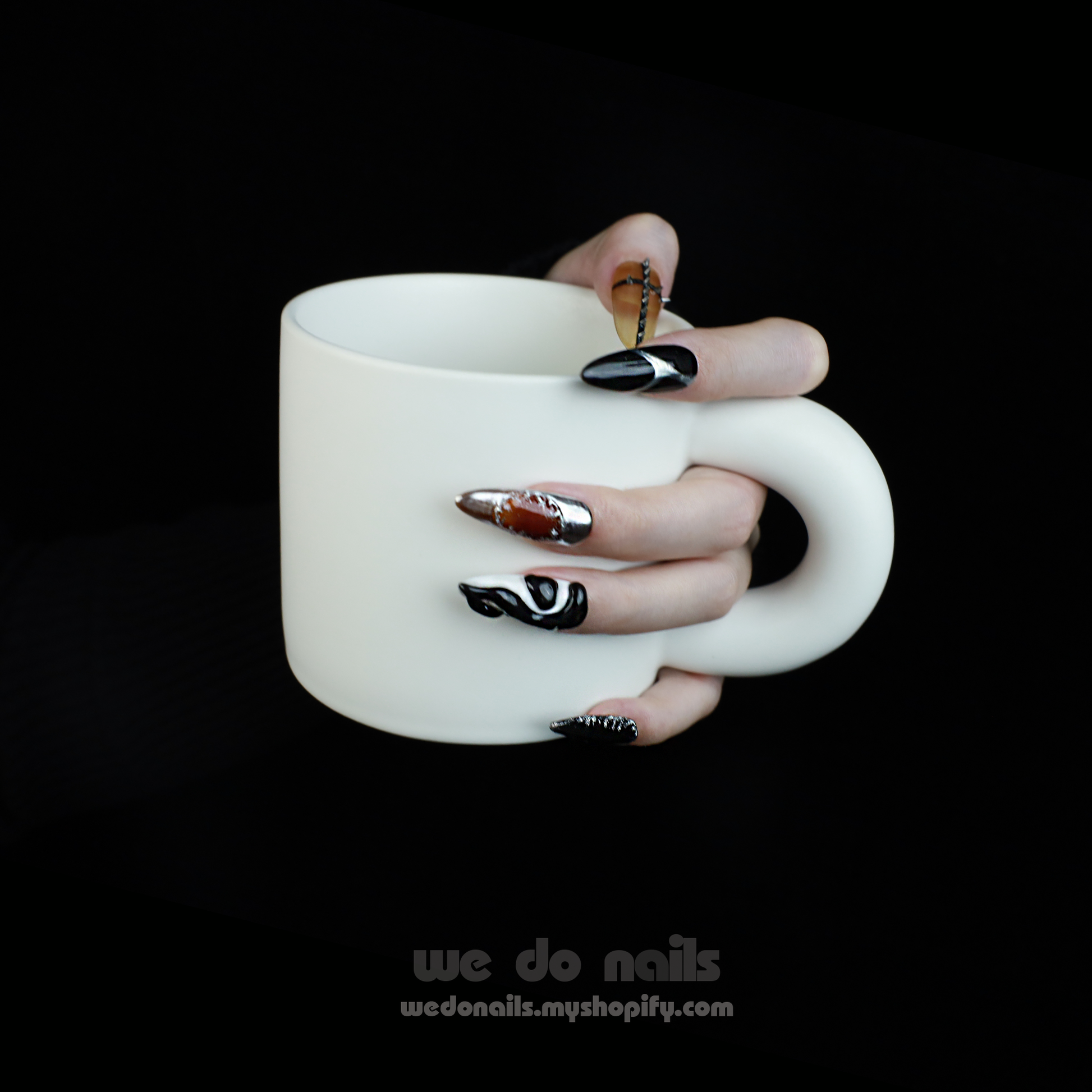 A hand holds a white mug, featuring long, intricately designed black and white Cool Rock Press-On Nails by We Do Nails from the Futuristic Noir Collection, adorned with metallic and gem accents against a dark background—an embodiment of avant-garde flair.