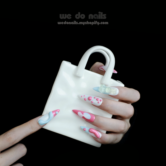 A close-up of a hand with intricate pink and blue 3D Rabbit Delight Press-On Nails by We Do Nails, holding a small white handbag against a black background. The nail design features swirls, dots, and raised elements, showcasing creative and colorful styling.
