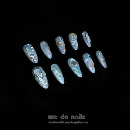 Displaying interlocked fingers adorned with We Do Nails Iridescent Bubble Press-On Nails, these long, pointed artificial nails feature a holographic silver and pearl finish, beautifully contrasted by a sleek black garment. Text reads we do nails and wedonails.myshopify.com.