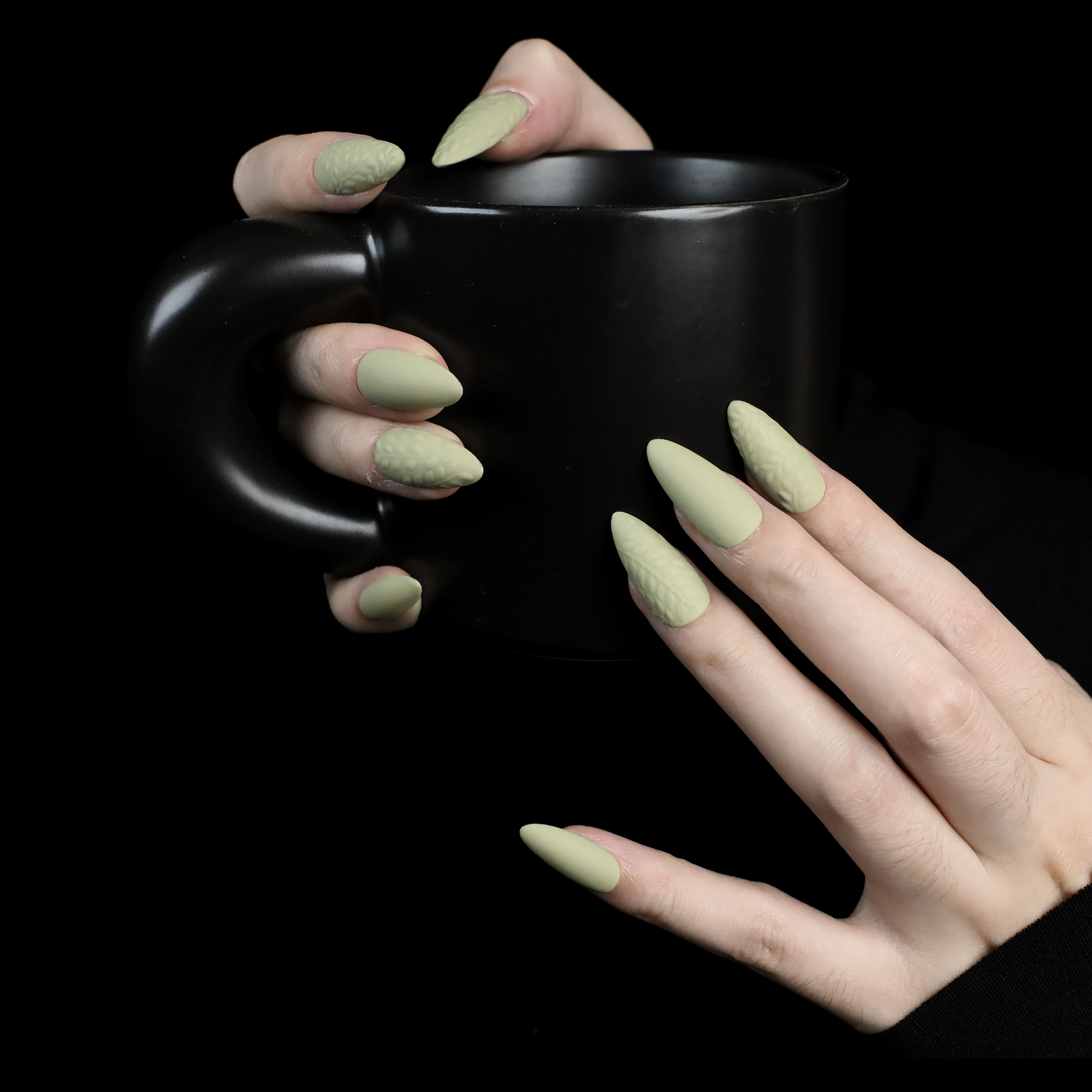 A person displays their crossed hands against a black background, showcasing We Do Nails Earthy Olive Green Press-On Nails. The textured olive design is elegantly highlighted by the sleeves of their black garment.