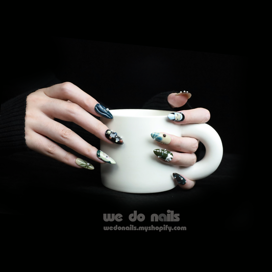 A person with long, artistic Tokyo Impression Press-On Nails by We Do Nails, featuring various designs and colors, holds a large white mug against a black background, highlighting the detailed nail art. Text reads We Do Nails with a website link.
