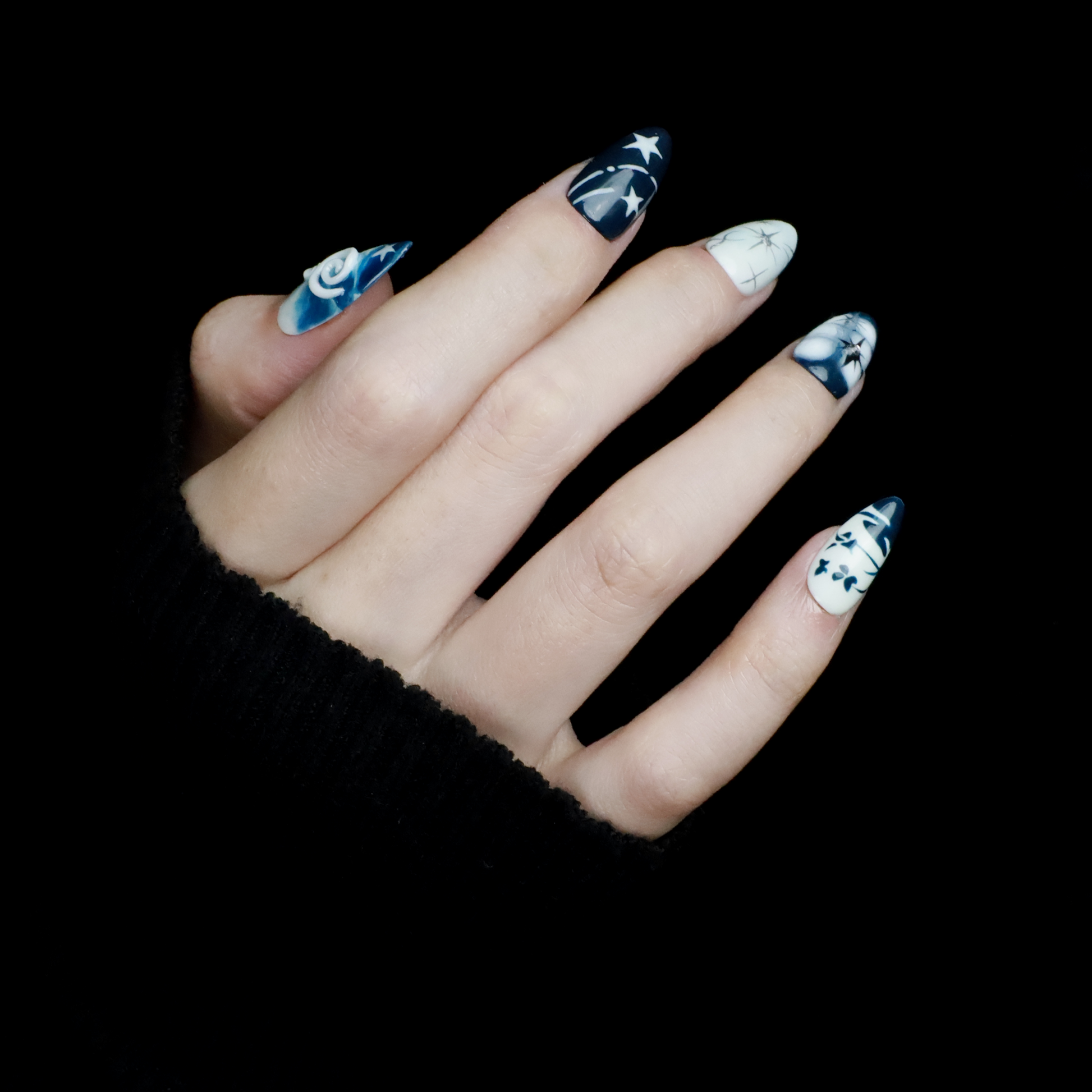 Hands adorned with We Do Nails Blue & White Porcelain Inspired Nails, showcasing long, almond-shaped designs with intricate traditional floral and abstract patterns. The nails stand out against black sleeves and a black background, reflecting the elegance of Chinese porcelain.