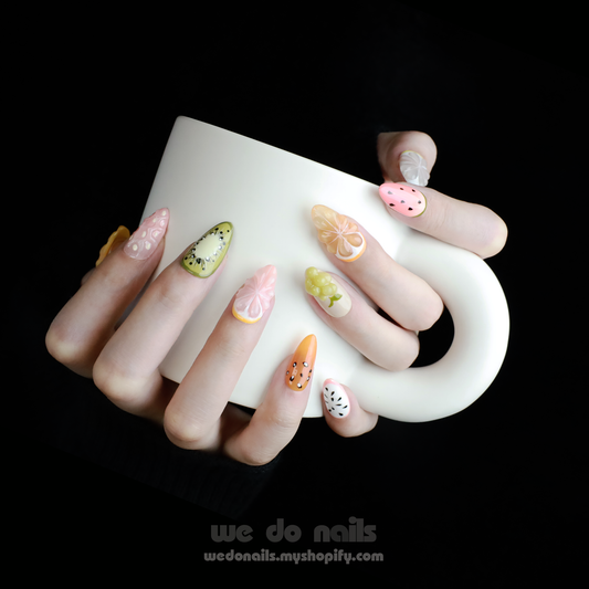 A hand holding a white mug displays vibrant, fruit-inspired press-on nails from We Do Nails Fruity Fantasy Collection, featuring kiwi, orange, strawberry, grape, and lemon designs on a sleek black background.