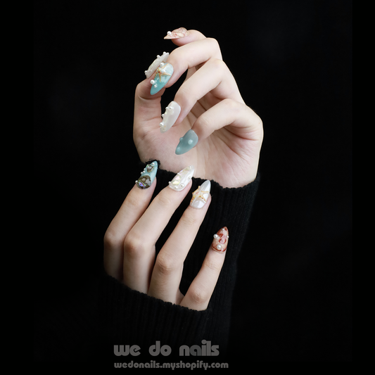 A hand with long, decorated Oceans Treasures Press-On Nails by We Do Nails holds one finger against a black background. The nails feature intricate 3D pearls and geometric patterns, adding elegance to the perfect summer look.