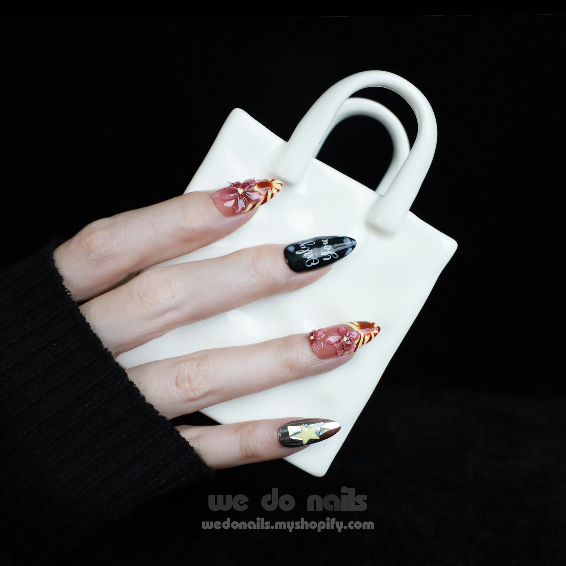 A hand adorned with We Do Nails Extravaganza Press-On Nails, showcasing unique patterns and colors, holds a small white handbag. The dazzling metallics gleam against the black background, highlighting the bold nail art and bag.