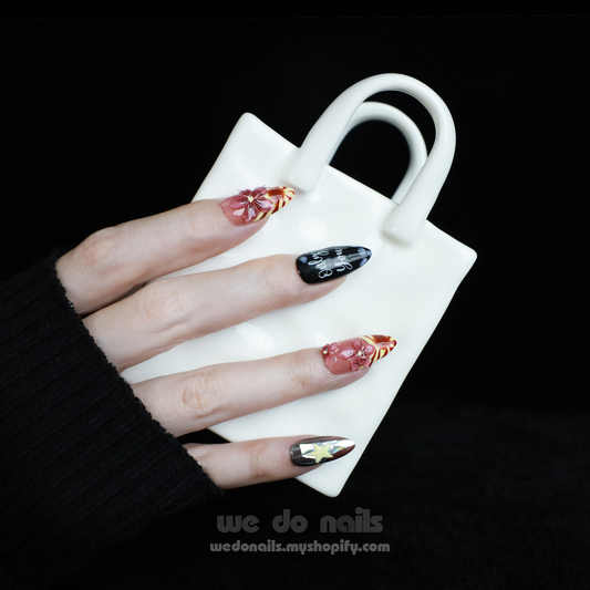 A hand adorned with We Do Nails Extravaganza Press-On Nails, showcasing unique patterns and colors, holds a small white handbag. The dazzling metallics gleam against the black background, highlighting the bold nail art and bag.