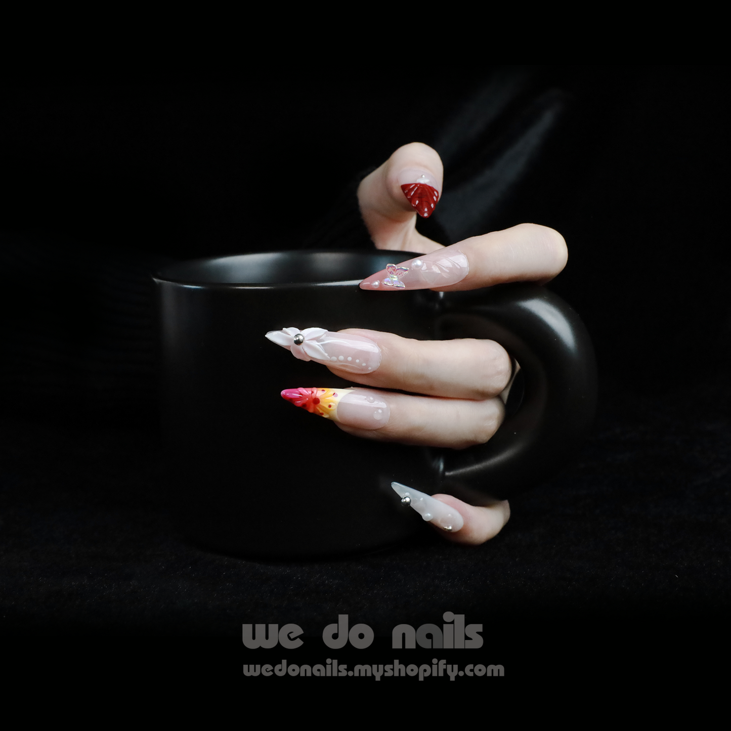 A hand flaunting Floral Fantasy Press-On Nails, accented with 3D flowers and rhinestones, grasps a large black mug. Against a dark backdrop, the foreground displays the text We Do Nails with their nail shop URL.