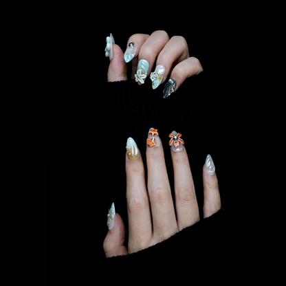 A hand with long, intricately decorated nails exhibits ethereal designs of blue and white swirls, 3D floral accents, pearls, and a metallic silver textured nail against a black background. This look perfectly captures the elegance of We Do Nails Celestial Blossom Press On Nails.