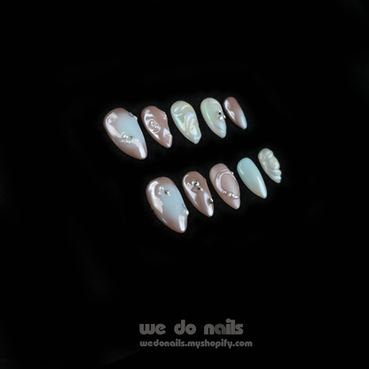 Close-up of hands with Pearl Dreams Press-On Nails by We Do Nails, showcasing manicured nails in small patterns and neutral colors. Subtle pearlescent hues add elegance. Fingers are gently intertwined, wearing a black long-sleeve sweater. We Do Nails text visible on the left.