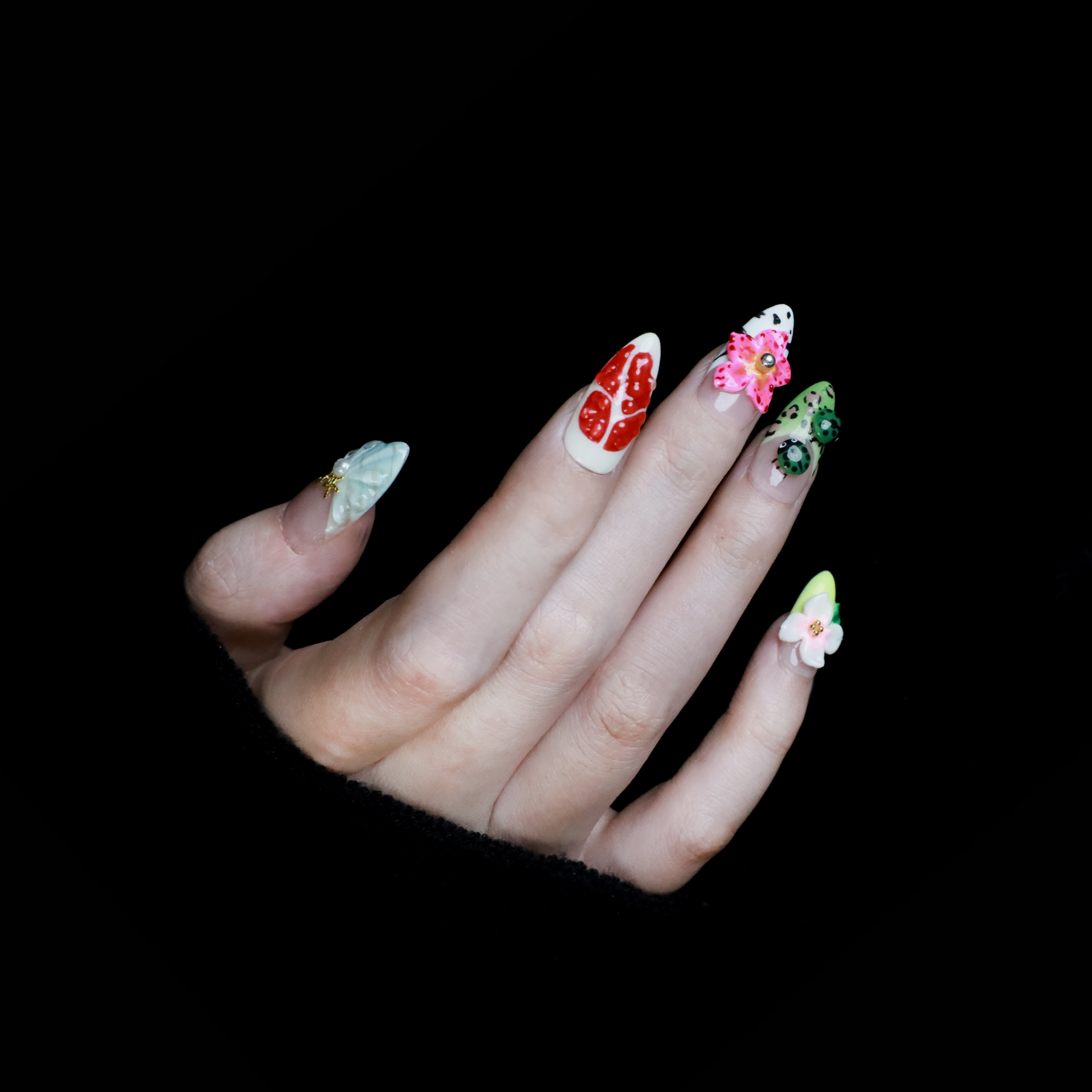 The Garden Blooms Press-On Nails by We Do Nails feature a whimsical garden theme with long, decorative nails, including a blue flower, cherries, red and white patterns, pearls, and stripes. Set against a black background, the hand is partially covered by a black sleeve.