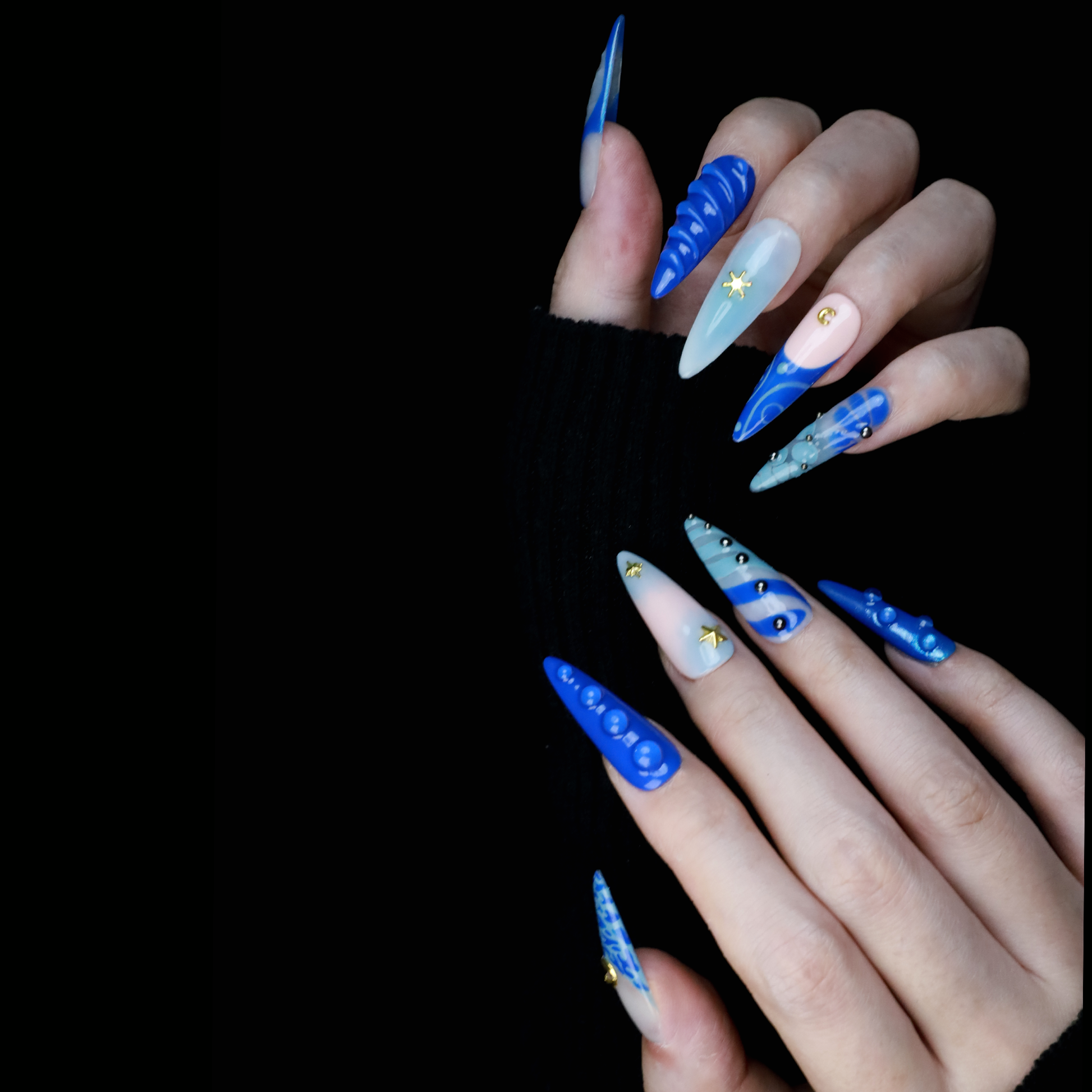 A hand featuring We Do Nails Blue Dreams Press-On Nails with elongated designs and pink hues, includes 3D metallic nail art and textured patterns like ridges and dots against a black backdrop.