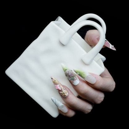 A hand flaunts We Do Nails Floral Blush Pearl Press-On Nails: green with pearls, pink with a butterfly, clear with gold accents, blue with a wave pattern, and a pink-white thumb—all set against a black background for an enchanting look.