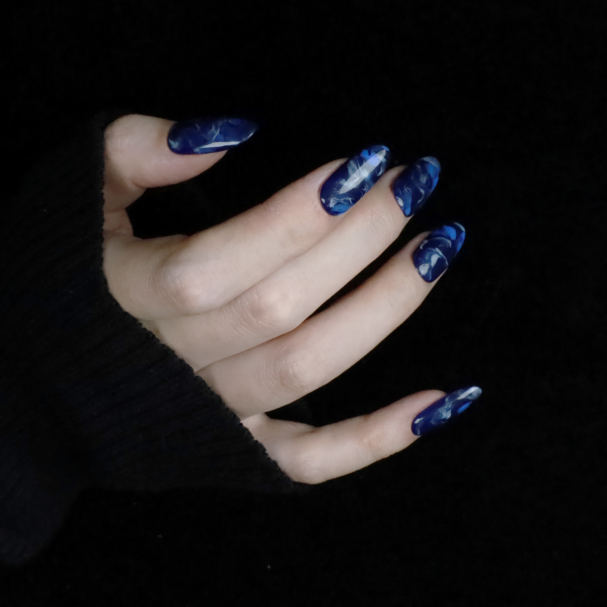 Hands display the Elegant Blue Marble Press-On Nails from We Do Nails, featuring glossy long oval shapes with a blue marble effect. A black ribbed sweater against a black background highlights these striking nails.