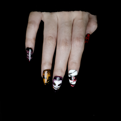 A close-up of two hands shows off the eerie Spooky Nightmares Halloween Press-On Nails by We Do Nails, featuring hand-painted designs like clowns, a pumpkin, and abstract art on a black background while holding a large white mug.