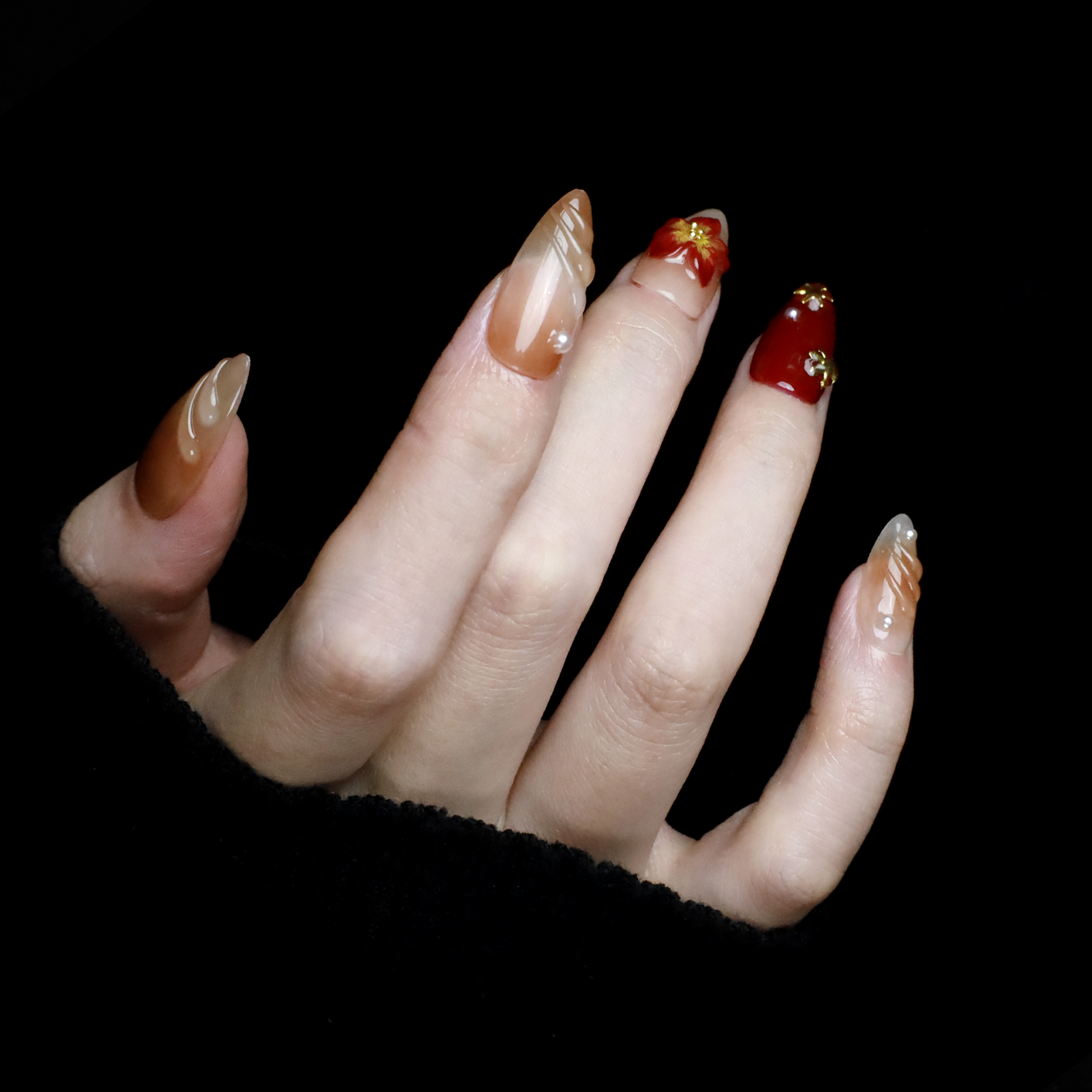 A pair of hands adorned with Elegant Floral Red and Beige Press-On Nails by We Do Nails, featuring intricate red flowers and pearl-gold accents, holds a large black mug. The elongated, almond-shaped nails boast a glossy finish with 3D floral designs against the black background.