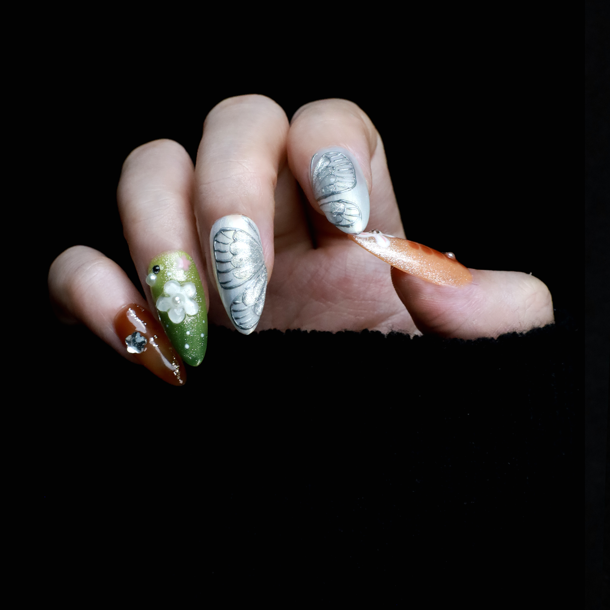 A hand showcases the Fantasy Garden Press-On Nails by We Do Nails against a black background. The nails feature designs of a koi fish with a pearl, peach with rhinestones, 3D green flowers, green with red floral dots, and metallic silver with a crystal accent.