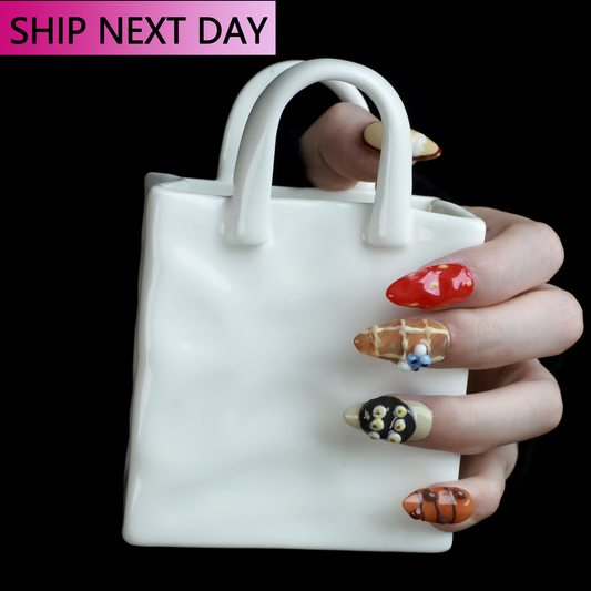 A hand with whimsical nail art holds a small white ceramic handbag against a black backdrop. SHIP NEXT DAY is displayed in pink at the top. The We Do Nails Gourmet Delight Press-On Nails promise vibrant patterns to dazzle for any occasion.