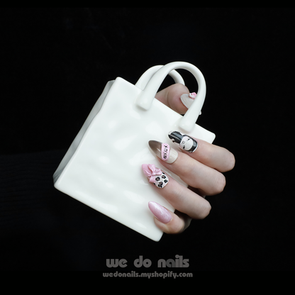 A stylish hand sports We Do Nails XOXO Feminine Press-On Nails with black, pink, and white designs. The nails feature cute patterns like a bow, a face, and OMG while elegantly holding a white ceramic handbag-shaped mug.