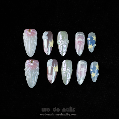 A hand with long, intricately designed Ocean-Themed Press-On Nails by We Do Nails holds a small white ceramic purse. The nails feature pastel floral and star marine motifs. A pink SHIP NEXT DAY banner appears in the top left corner.