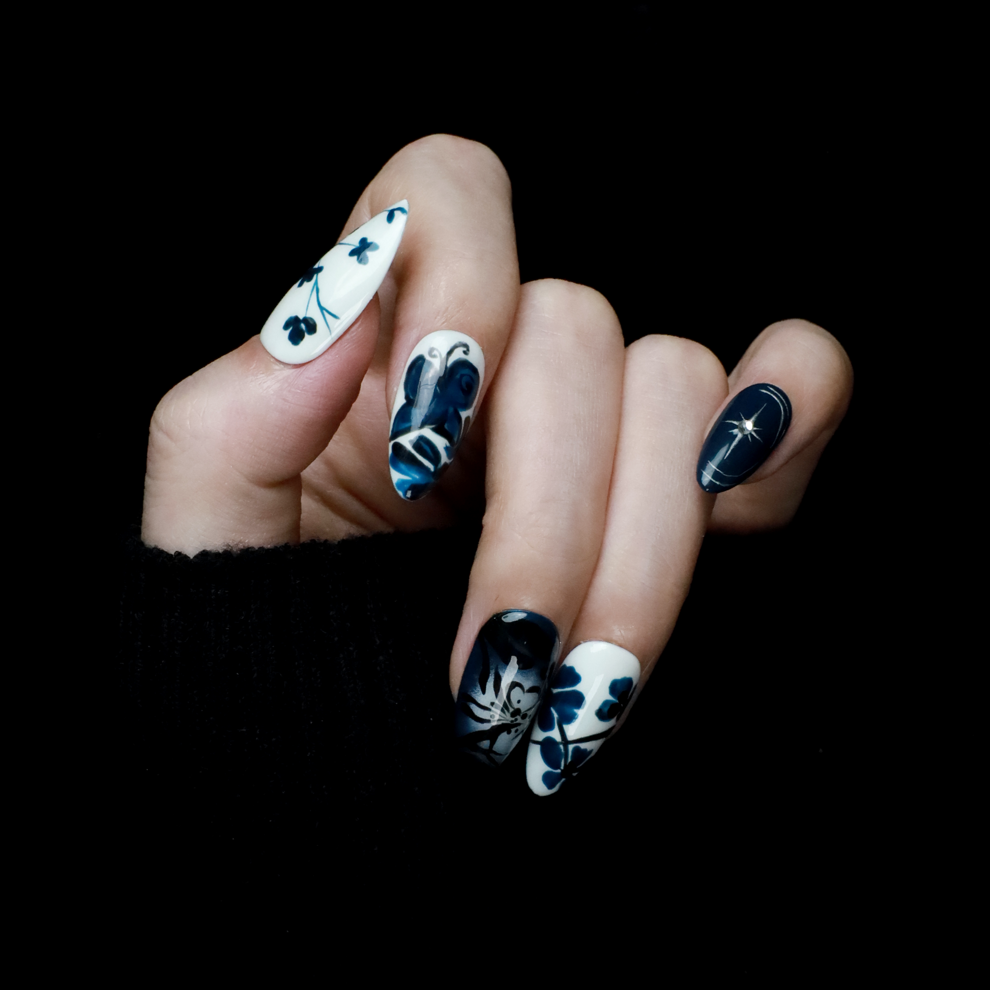 Hands adorned with We Do Nails Blue & White Porcelain Inspired Nails, showcasing long, almond-shaped designs with intricate traditional floral and abstract patterns. The nails stand out against black sleeves and a black background, reflecting the elegance of Chinese porcelain.