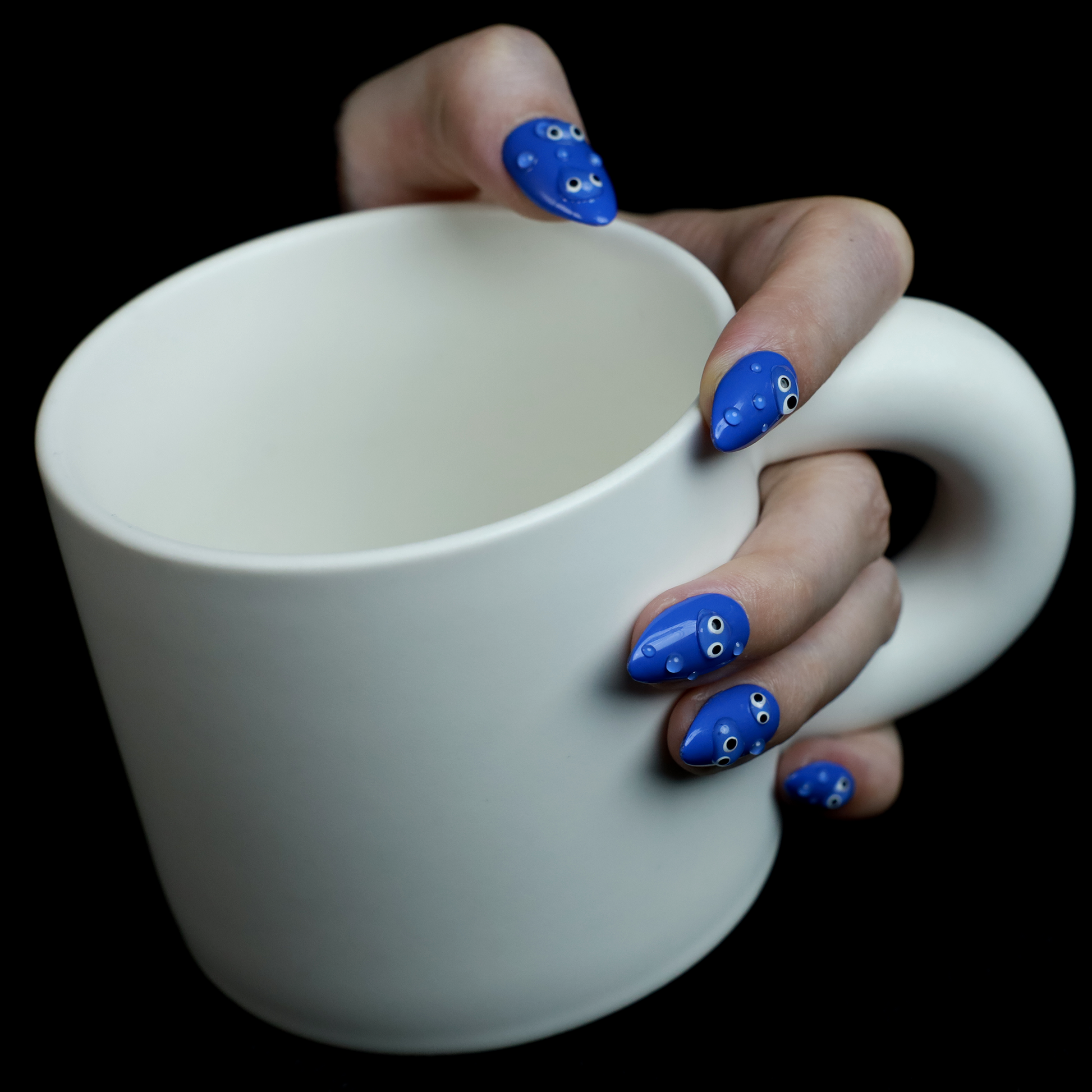 A hand with We Do Nails Cute Blue Monster Press-On Nails set features long, almond-shaped, bright blue nails adorned with playful cartoon faces and 3D googly eyes. The hand in a black sweater is striking against the dark background.