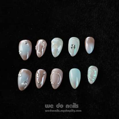 Close-up of hands with Pearl Dreams Press-On Nails by We Do Nails, showcasing manicured nails in small patterns and neutral colors. Subtle pearlescent hues add elegance. Fingers are gently intertwined, wearing a black long-sleeve sweater. We Do Nails text visible on the left.