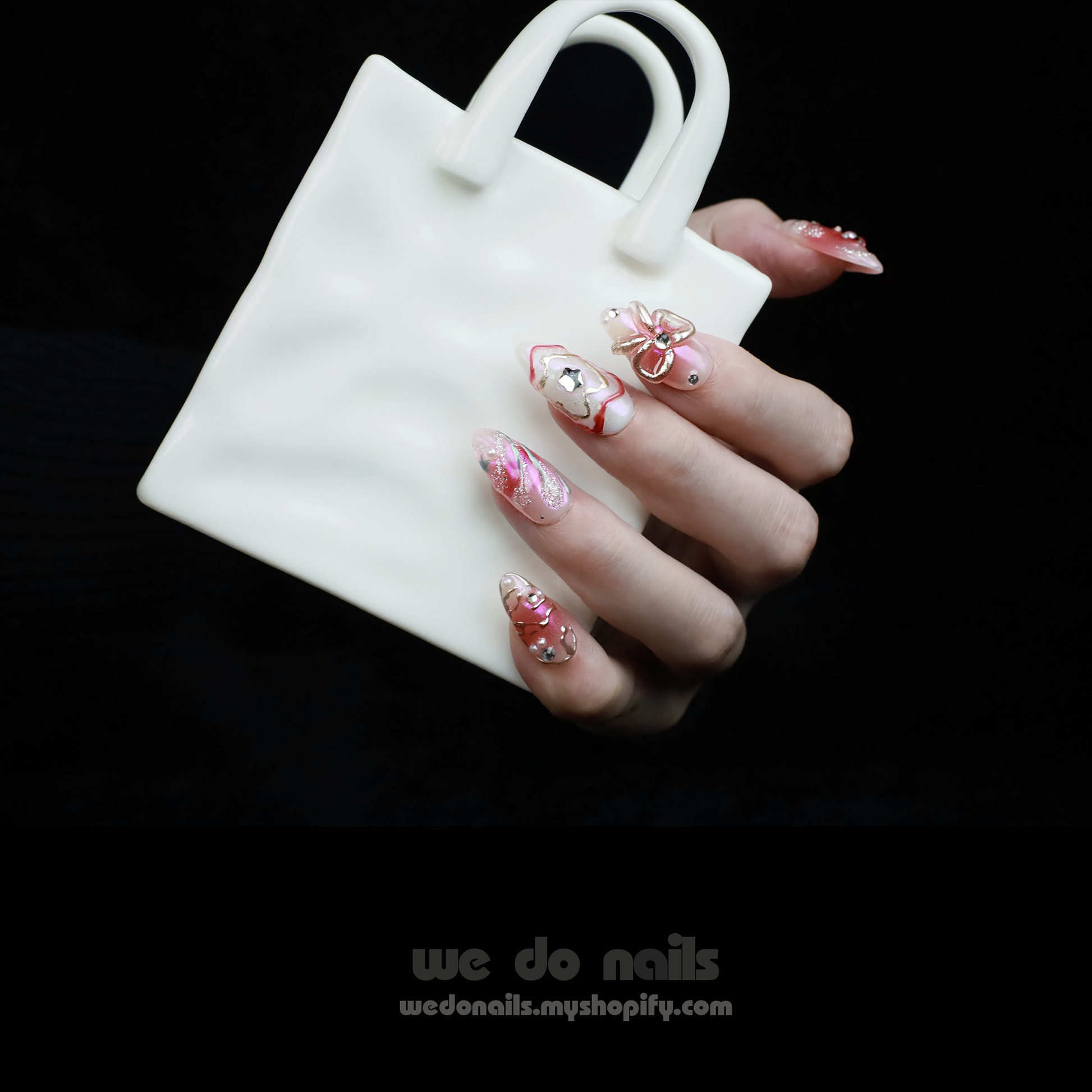 A person with intricately designed Blushing Charm Press-On Nails by We Do Nails holds a small, white ceramic handbag. The nails display soft pinks and abstract patterns in pink, white, and gold, accented with shimmering details and decorative embellishments.