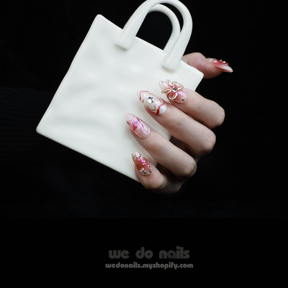 A person with intricately designed Blushing Charm Press-On Nails by We Do Nails holds a small, white ceramic handbag. The nails display soft pinks and abstract patterns in pink, white, and gold, accented with shimmering details and decorative embellishments.
