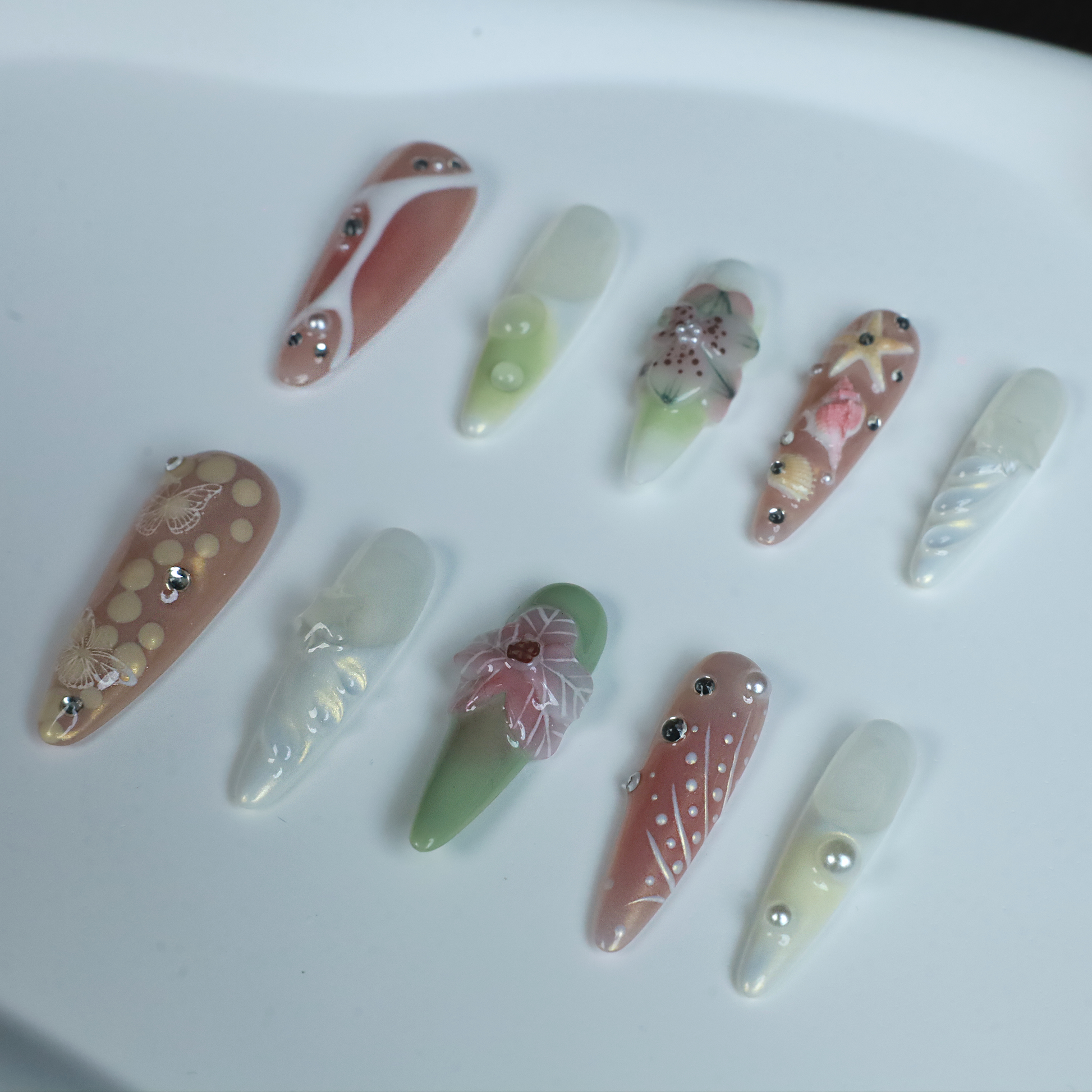 A hand flaunts We Do Nails Floral Blush Pearl Press-On Nails: green with pearls, pink with a butterfly, clear with gold accents, blue with a wave pattern, and a pink-white thumb—all set against a black background for an enchanting look.