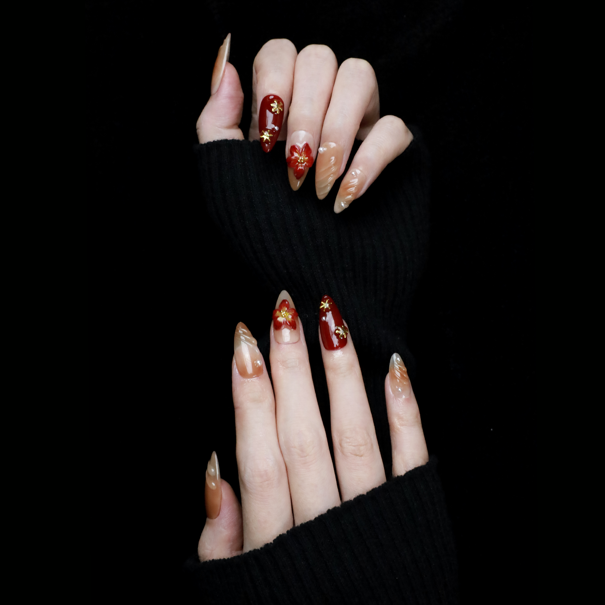 A pair of hands adorned with Elegant Floral Red and Beige Press-On Nails by We Do Nails, featuring intricate red flowers and pearl-gold accents, holds a large black mug. The elongated, almond-shaped nails boast a glossy finish with 3D floral designs against the black background.