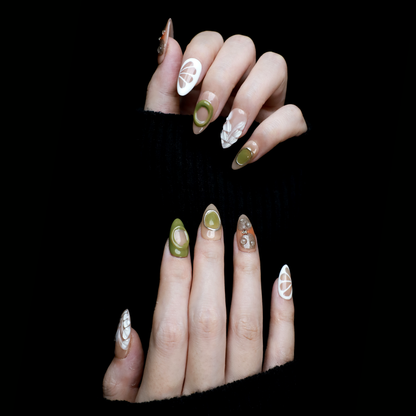 A hand in a black sleeve displays the Leaf and Organic design Press-On Nails by We Do Nails, featuring long acrylic nails with white abstract patterns, green accents, and clear 3D embellishments that create a nature-inspired look against the dark background.