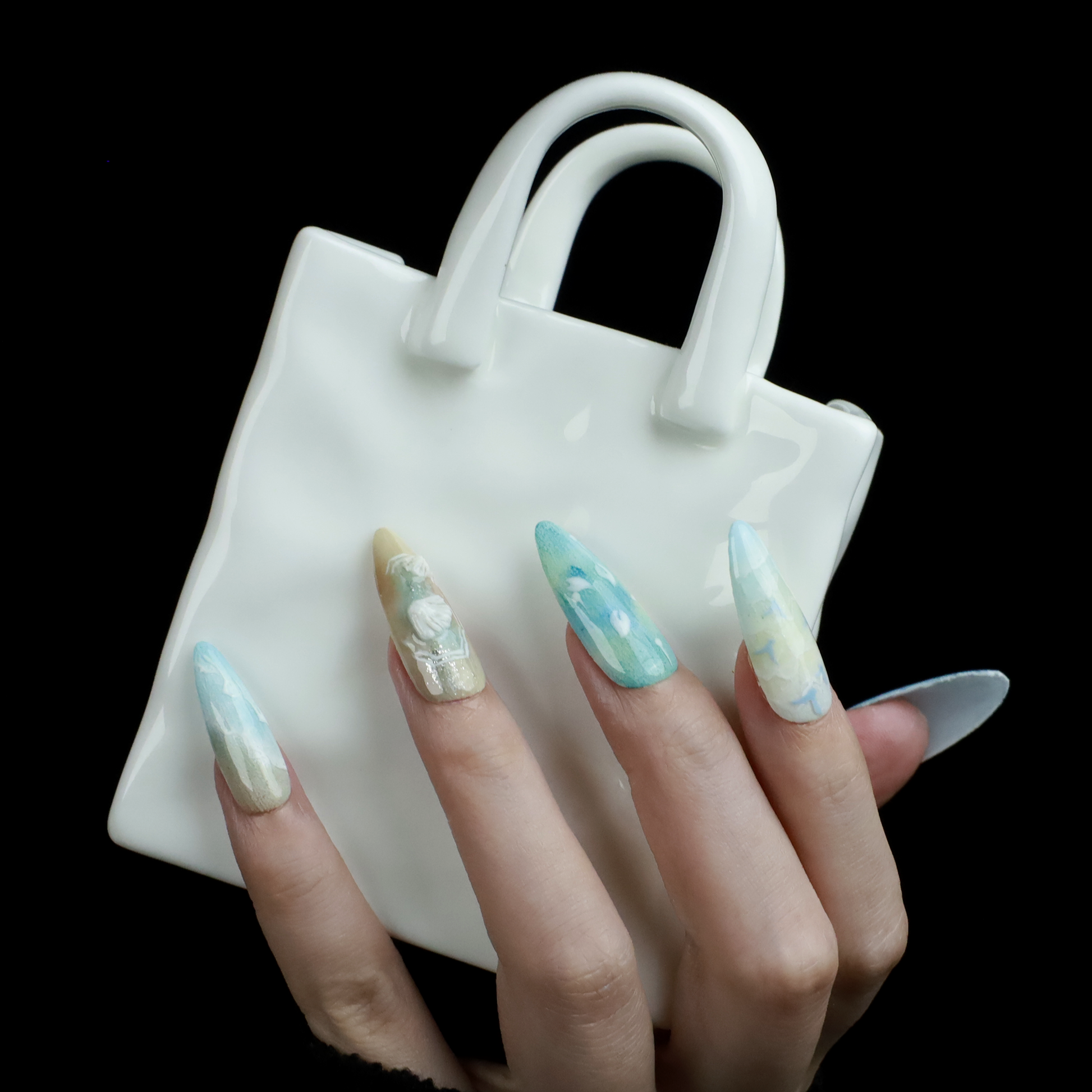 A hand flaunts the elegant Ocean-Inspired Press-On Nails by We Do Nails, featuring long, pointed shapes with ocean-themed designs. The set showcases ocean breeze colors, seashell art, and wave patterns against a dark background to highlight its intricate beauty.