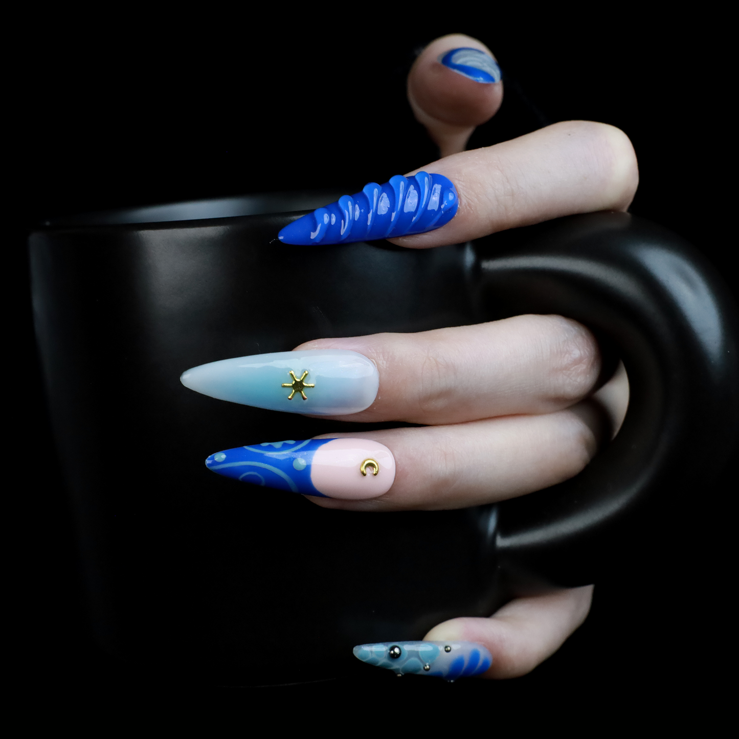 A hand featuring We Do Nails Blue Dreams Press-On Nails with elongated designs and pink hues, includes 3D metallic nail art and textured patterns like ridges and dots against a black backdrop.