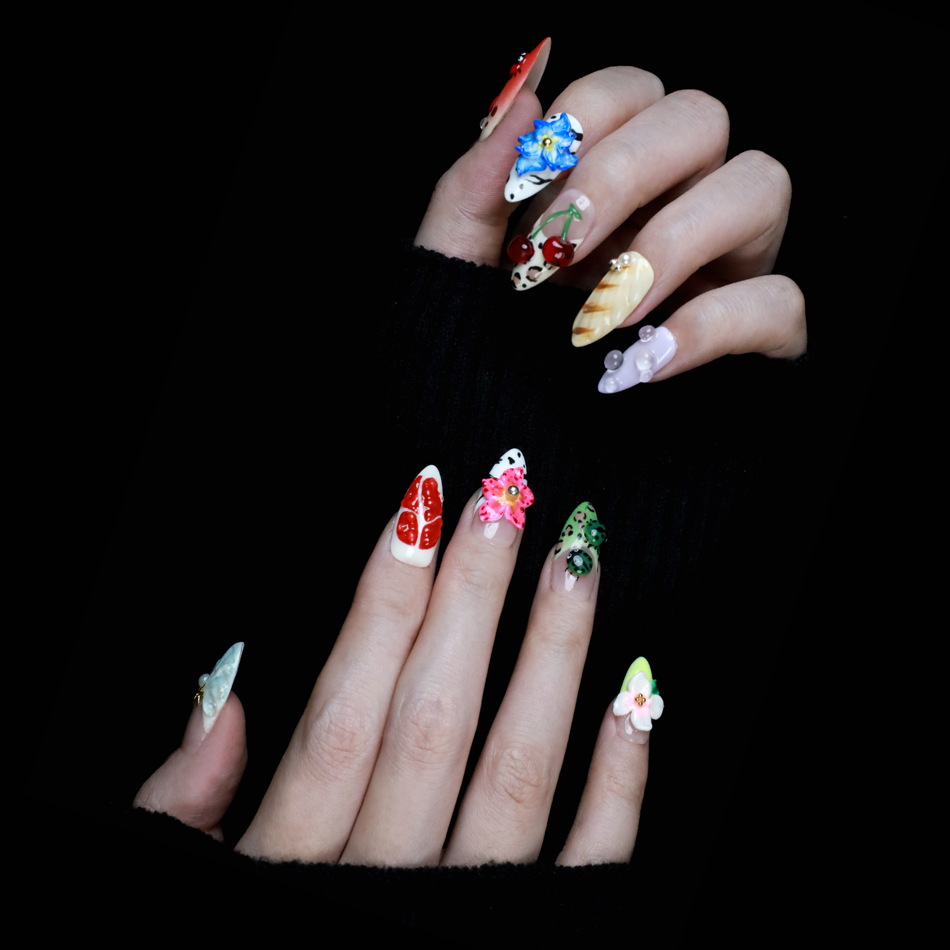 The Garden Blooms Press-On Nails by We Do Nails feature a whimsical garden theme with long, decorative nails, including a blue flower, cherries, red and white patterns, pearls, and stripes. Set against a black background, the hand is partially covered by a black sleeve.