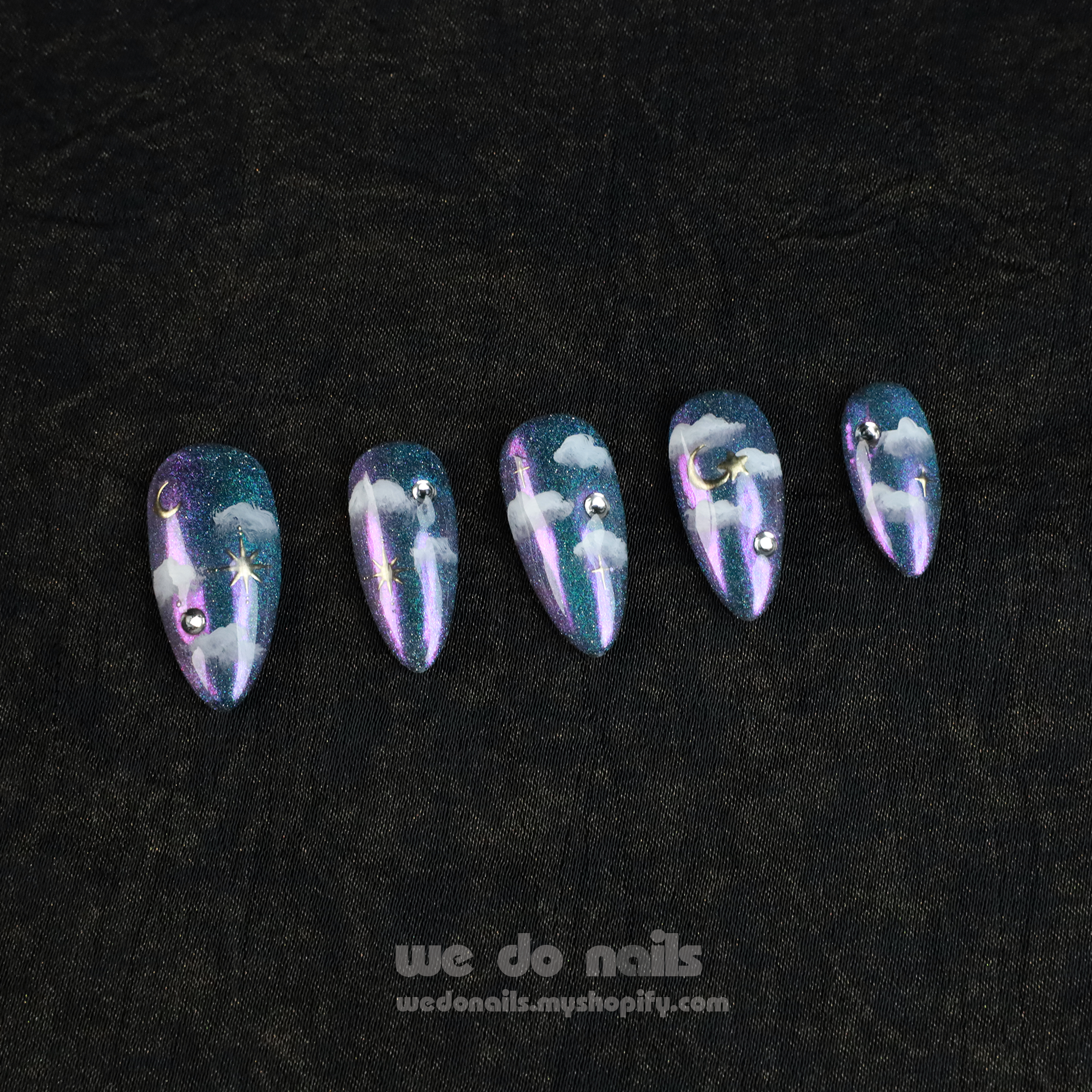 A hand showcases Starry Night Press-On Nails with long, glittery tips featuring a cloud and starry night design adorned with small gems. The person wears a black sleeve. Text below reads: We Do Nails with a website link.