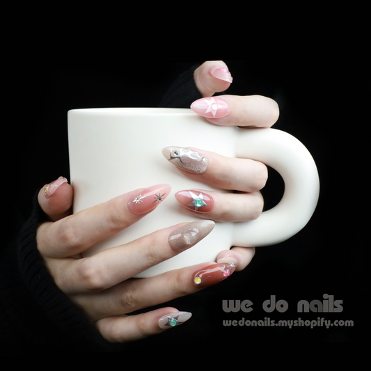 Hands hold a white mug, displaying Shiny Star Press-On Nails by We Do Nails. Each celestial-inspired, pastel design glitters with stars. Set against a black sweater, We Do Nails is written in the corner, guaranteeing salon-quality manicure perfection.