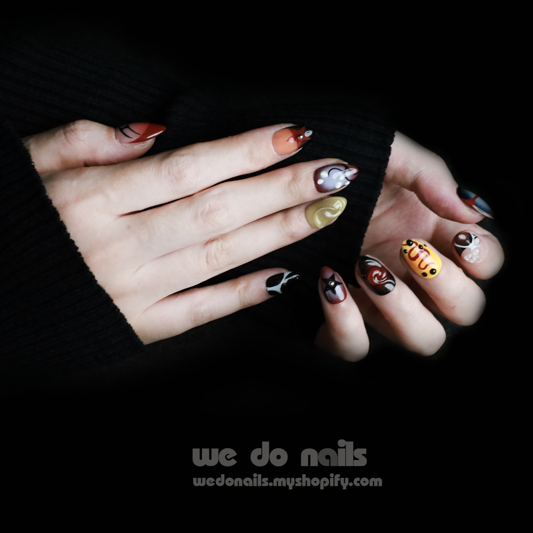 A hand with trendy Snake-inspired Press-On Nails features a yellow smiley face, black and white patterns, and a butterfly. Set against a black background, We Do Nails appears with a URL below.