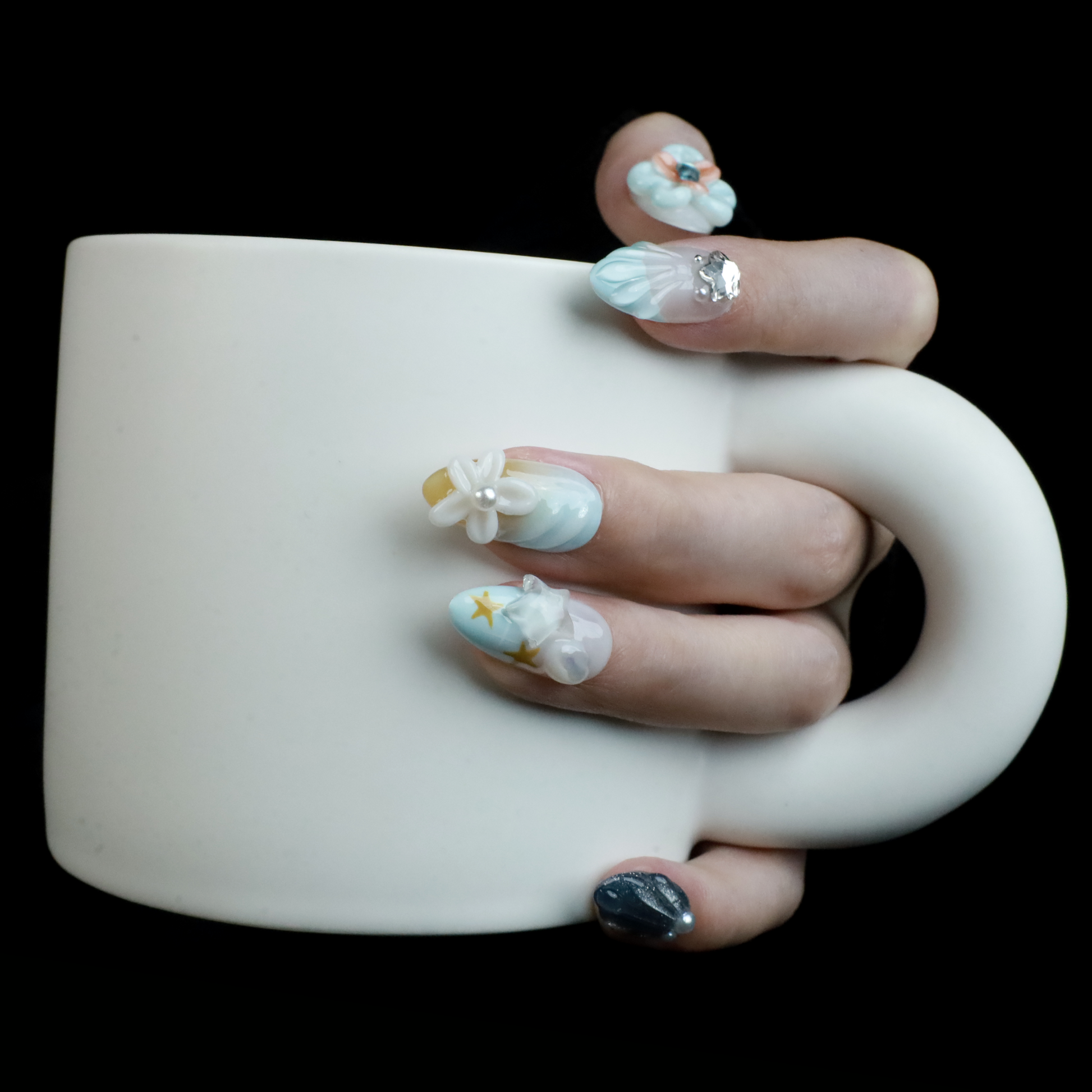 A hand with long, intricately decorated nails exhibits ethereal designs of blue and white swirls, 3D floral accents, pearls, and a metallic silver textured nail against a black background. This look perfectly captures the elegance of We Do Nails Celestial Blossom Press On Nails.