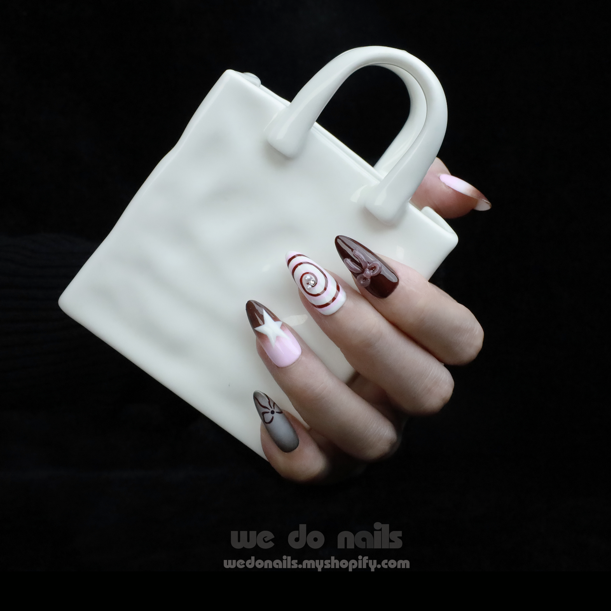 A hand with enchanting nails holds a small white handbag. The Charming Cocoa Delight Press-On Nails from We Do Nails showcase intricate patterns, featuring stripes and dotted embellishments in white, brown, and red shades, all set against a black background that enhances their allure.