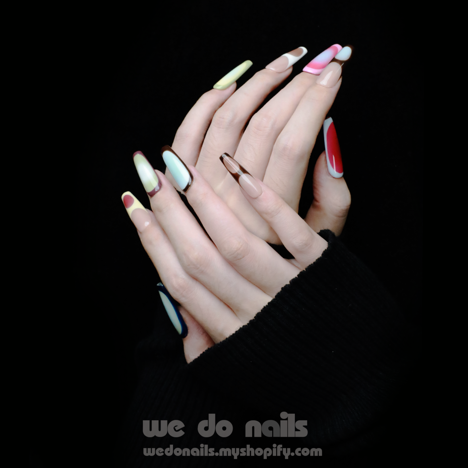 A pair of hands with long, artistic Ms.Circles Press-On Nails by We Do Nails stand out against a black background. The vibrant nails have bold patterns in green, pink, and nude hues. Partially emerging from black sweater sleeves, they create a striking contrast.