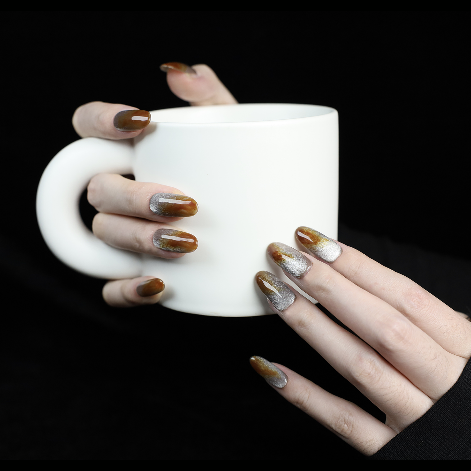 A hand with We Do Nails Elegant Brown Marble Press-On Nails, featuring a subtle metallic chrome accent, holds a simple white mug against a black backdrop.