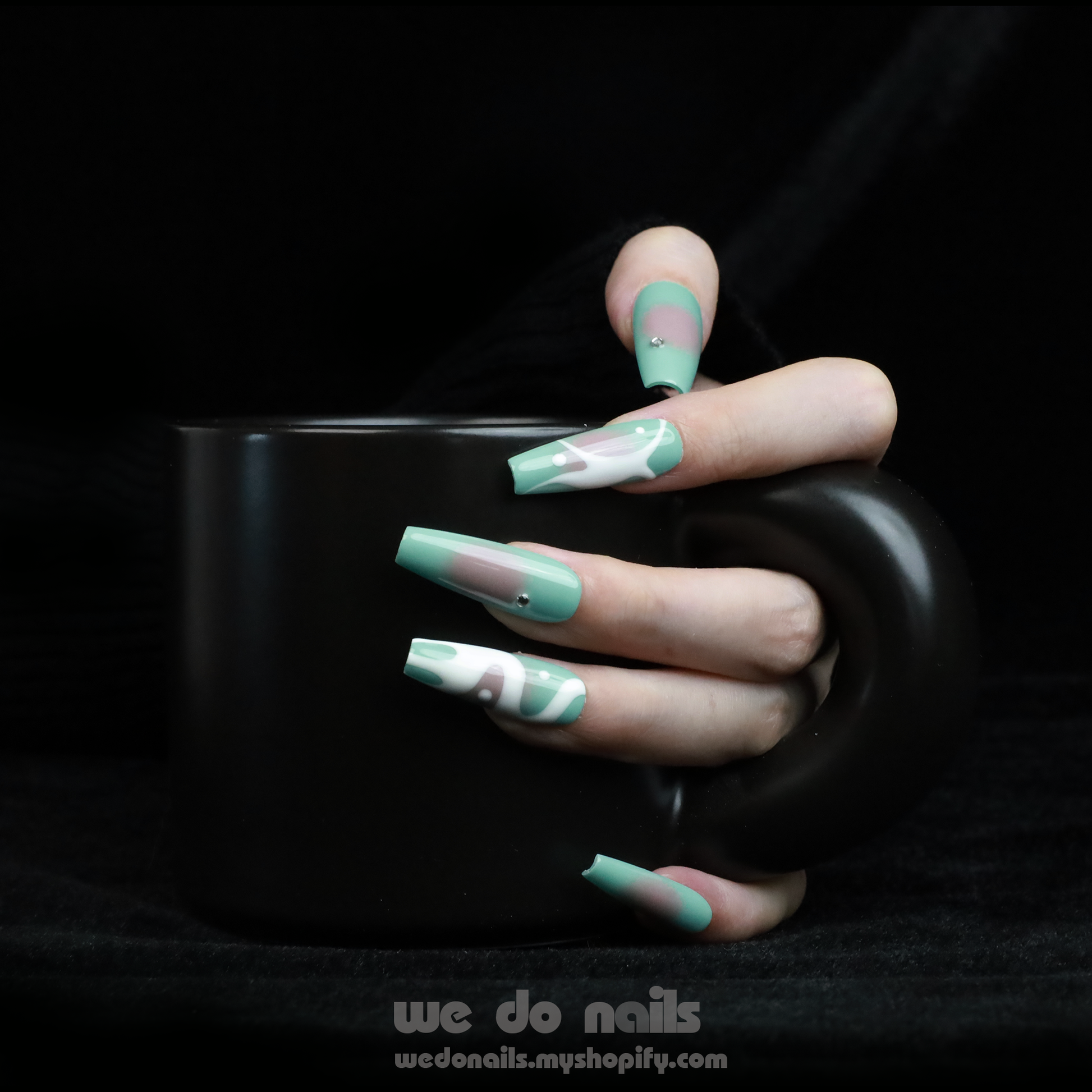 Hands with long, angular green nails pose gracefully against a black background, showcasing a modern and stylish design. The glossy finish and rings add flair, elegantly highlighting the allure of Spilled Milk Press-On Nails by We Do Nails.