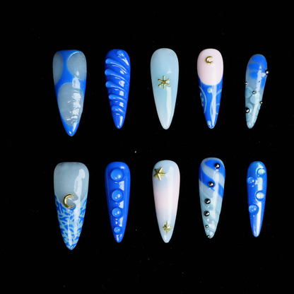 A hand featuring We Do Nails Blue Dreams Press-On Nails with elongated designs and pink hues, includes 3D metallic nail art and textured patterns like ridges and dots against a black backdrop.
