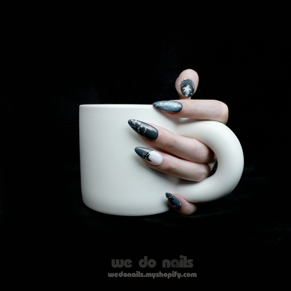 A person holds a white mug with both hands, showcasing long, pointed nails from We Do Nails Midnight Custom Press-On Nails. Star and galaxy designs exude cosmic elegance against a dark background, enhancing the vibrant celestial wonder of the nail art.