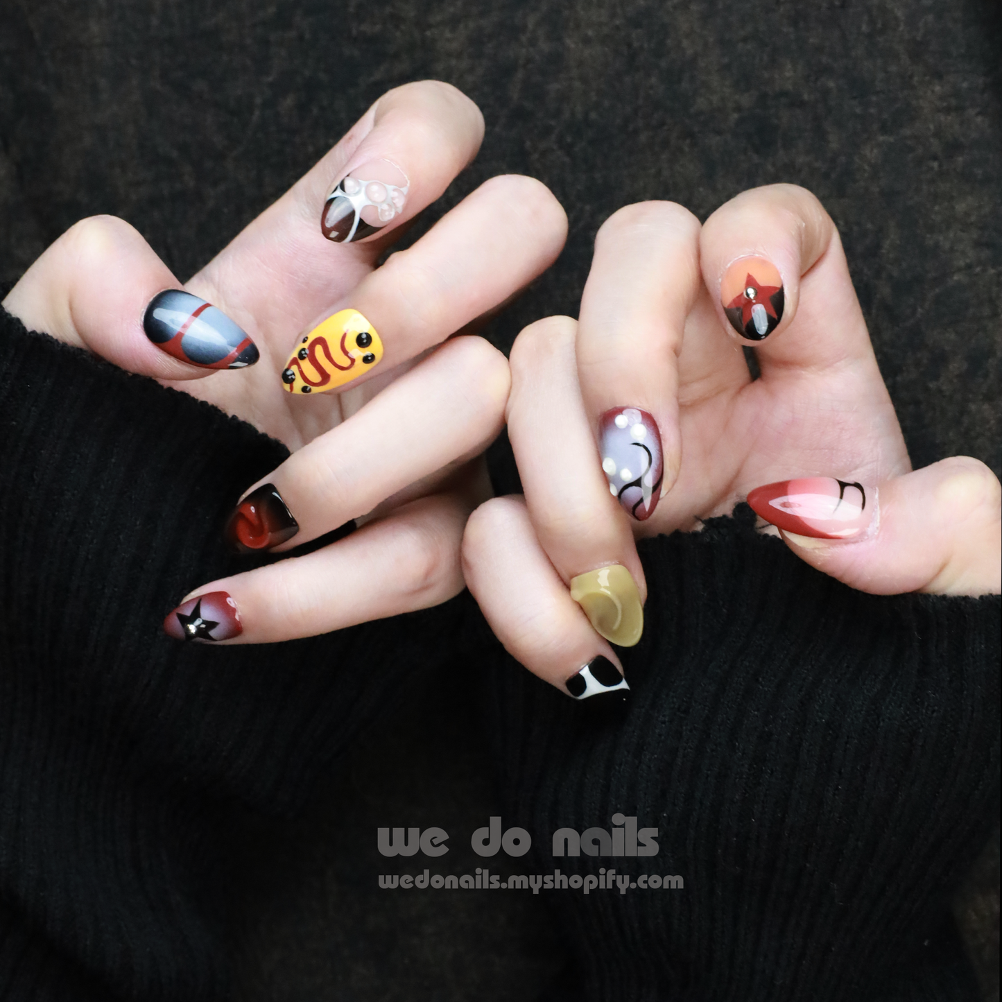 A hand with trendy Snake-inspired Press-On Nails features a yellow smiley face, black and white patterns, and a butterfly. Set against a black background, We Do Nails appears with a URL below.